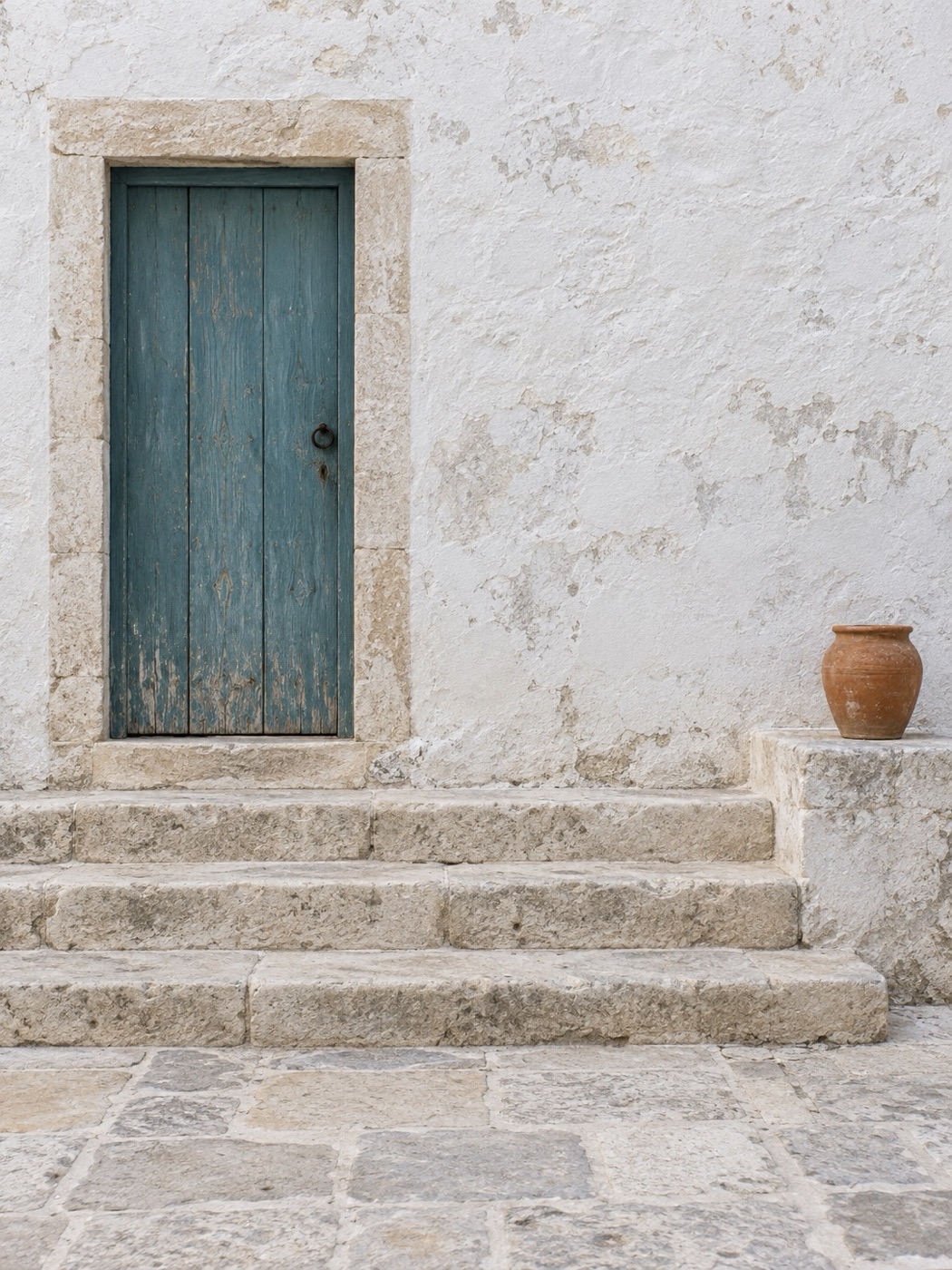 An empty Mediterranean village lane: a weathered teal wooden door in a stone frame, three worn stone steps, and a terracotta pot on a low stone ledge